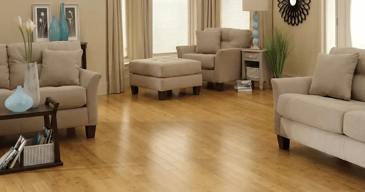 A living room with professionally installed bamboo flooring, beige sofas, a matching ottoman, a side table with a blue lamp, vases, framed pictures, and large windows with blinds and curtains in Louisville, KY